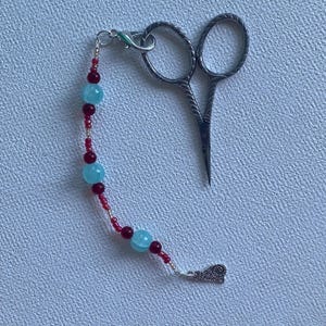 Può includere: Piccole forbici da ricamo color argento con un ciondolo di perline. Il ciondolo presenta perline azzurre e rosso scuro infilate su una catena d'argento. Le forbici hanno manici curvi e lame appuntite, ideali per lavori di cucito dettagliati.