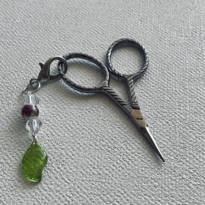 Può includere: Piccole forbici da ricamo argentate con un design decorativo a manico ritorto. Le forbici hanno un ciondolo con perline trasparenti e viola e un ciondolo a forma di foglia verde. Le forbici sono lunghe circa 10 cm.