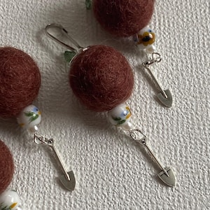 May include: Close-up of handmade earrings featuring maroon felt balls, floral beads, and miniature silver shovel charms. The earrings have a whimsical, garden-inspired theme, with a clip-on closure. The beads have a white base with blue and yellow floral designs.