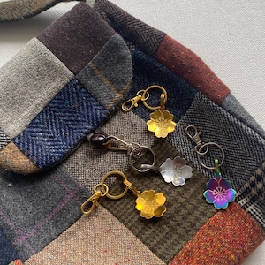 Puede incluir: Una colección de llaveros con colgantes en forma de flor en acabados dorados, plateados e iridiscentes. Los llaveros se muestran sobre un fondo de tela de retazos con varias texturas y colores, incluyendo marrón, azul y gris.