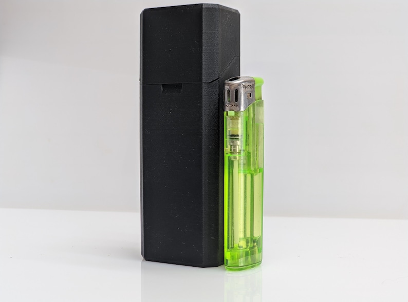 Puede incluir: Una funda rectangular negra est&aacute; junto a un encendedor verde transparente. La funda es alta y tiene un acabado mate. El encendedor es de tama&ntilde;o est&aacute;ndar, con una parte superior de metal y una c&aacute;mara de combustible visible.