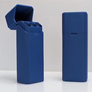 Puede incluir: Dos estuches rectangulares azul cobalto. Un estuche est&aacute; abierto, revelando tres compartimentos cil&iacute;ndricos. El otro estuche est&aacute; cerrado. Ambos estuches tienen una superficie texturizada y un dise&ntilde;o geom&eacute;trico. Los estuches est&aacute;n sobre una superficie blanca.
