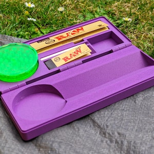 Può includere: Un vassoio per rollare viola con un grinder verde, cartine e filtri. Il vassoio è aperto, rivelando scomparti per accessori. Il logo del marchio RAW è visibile.
