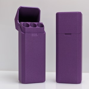 Puede incluir: Dos estuches rectangulares morados, uno abierto y otro cerrado, se muestran sobre un fondo blanco. El estuche abierto revela tres compartimentos cil&iacute;ndricos. Ambos estuches tienen una superficie texturizada y un dise&ntilde;o geom&eacute;trico.