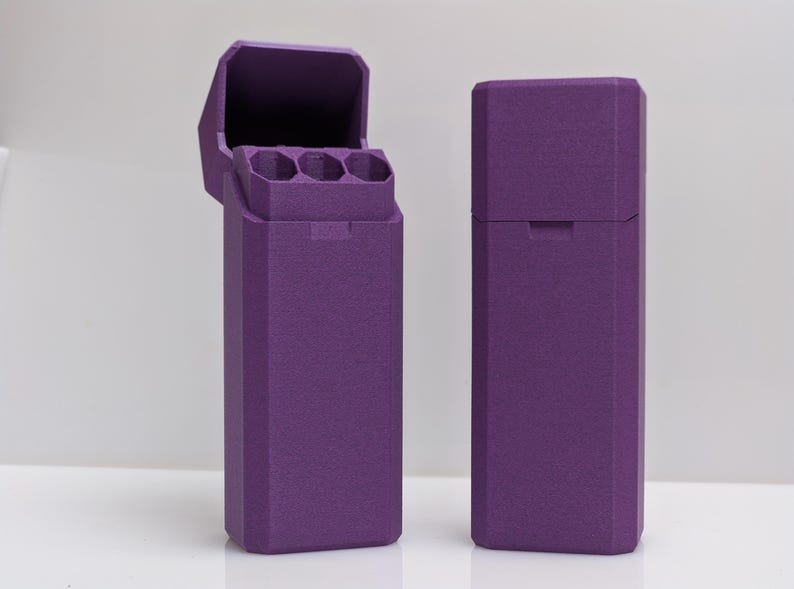 Puede incluir: Dos estuches rectangulares morados, uno abierto y otro cerrado, se muestran sobre un fondo blanco. El estuche abierto revela tres compartimentos cil&iacute;ndricos. Ambos estuches tienen una superficie texturizada y un dise&ntilde;o geom&eacute;trico.