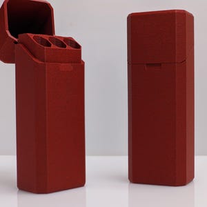 Puede incluir: Se muestran dos estuches rectangulares rojos, uno abierto y otro cerrado, sobre un fondo blanco. El estuche abierto revela tres compartimentos cil&iacute;ndricos. Los estuches tienen una superficie texturizada y una tapa con bisagras.