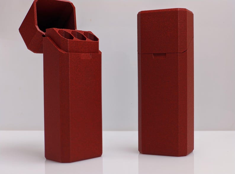 Puede incluir: Se muestran dos estuches rectangulares rojos, uno abierto y otro cerrado, sobre un fondo blanco. El estuche abierto revela tres compartimentos cil&iacute;ndricos. Los estuches tienen una superficie texturizada y una tapa con bisagras.