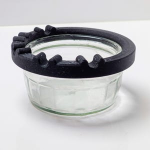 Puede incluir: Un tarro de cristal transparente con un borde negro texturizado que presenta una serie de muescas curvas. El tarro está sobre una superficie blanca. El borde parece estar hecho de un material impreso en 3D.