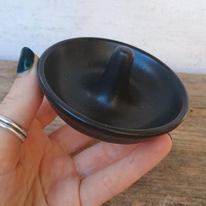 May include: A matte black ceramic ring dish featuring a circular base and a central conical post. The dish has a smooth, uniform finish and a shallow, bowl-like shape designed to hold jewelry items securely in place on a flat surface.