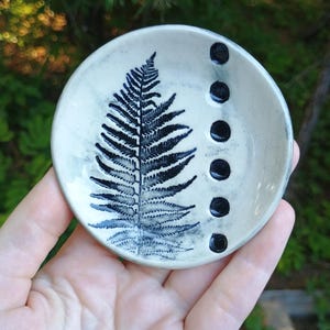 May include: A small, round ceramic dish with a fern leaf design in black and white. The dish has a light beige background and four black dots along the rim. The dish is held in a hand.