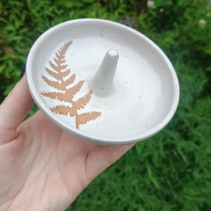 Puede incluir: Un plato de cer&aacute;mica redondo para anillos con un esmalte blanquecino moteado y un poste c&oacute;nico central. La superficie interior presenta una impresi&oacute;n de hoja de helecho marr&oacute;n. El plato tiene un borde liso y curvo y una base poco profunda.