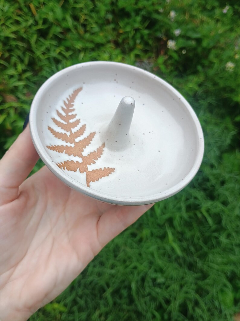 Puede incluir: Un plato de cer&aacute;mica redondo para anillos con un esmalte blanquecino moteado y un poste c&oacute;nico central. La superficie interior presenta una impresi&oacute;n de hoja de helecho marr&oacute;n. El plato tiene un borde liso y curvo y una base poco profunda.