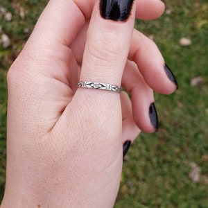 May include: A silver ring with a floral design on the band. The ring is worn on the thumb of a hand with black nail polish.