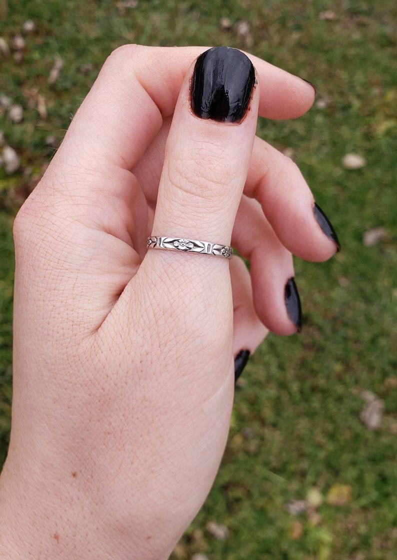 May include: A silver ring with a floral design on the band. The ring is worn on the thumb of a hand with black nail polish.