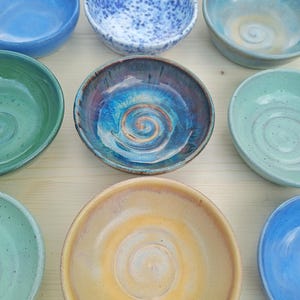 May include: A collection of eight small ceramic bowls in various colors including blue, green, yellow, and white with speckled or swirled glaze patterns. Each bowl features a distinct circular spiral design at the base of the interior.