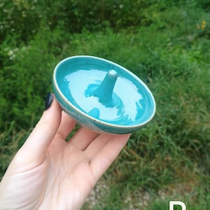 Puede incluir: Un plato de cer&aacute;mica redondo para anillos con un interior turquesa brillante y un cono central para sujetar joyas. El exterior presenta un acabado moteado con una base blanca, y la letra P es visible en la esquina inferior derecha.