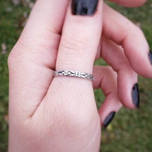 May include: A silver ring with a delicate floral design. The ring is worn on the index finger of a hand with black nail polish.