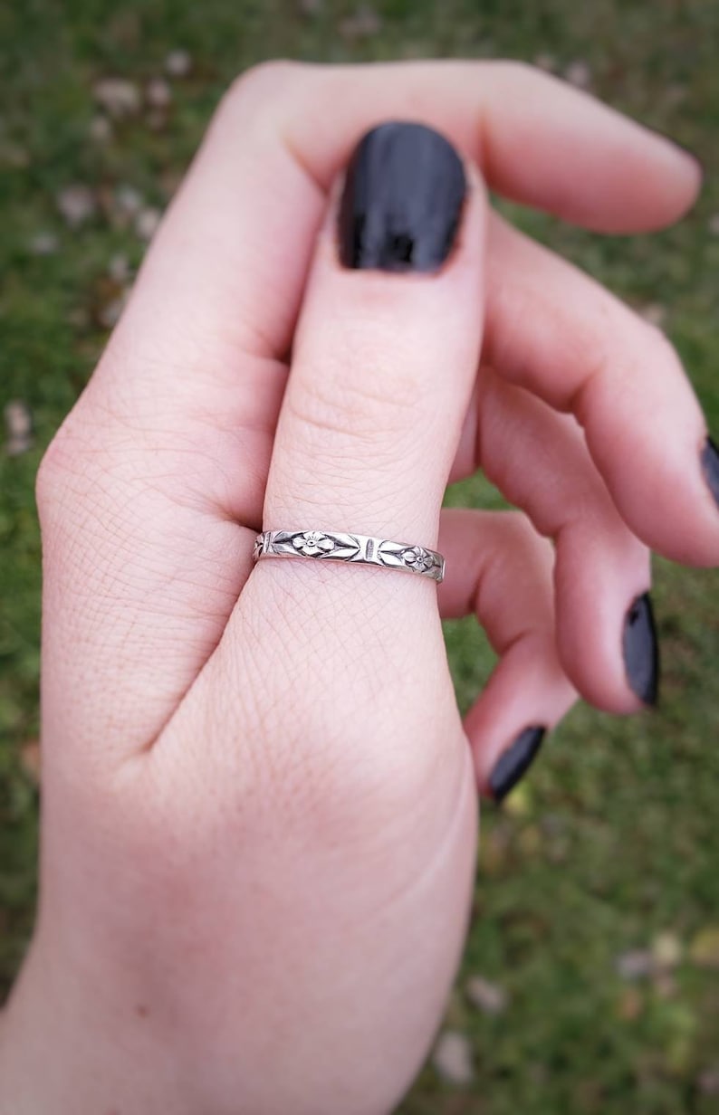 May include: A silver ring with a delicate floral design. The ring is worn on the index finger of a hand with black nail polish.