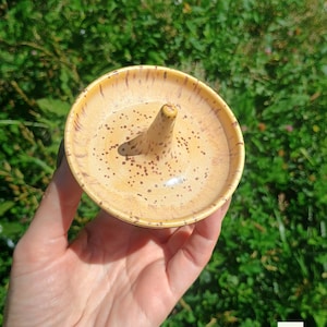 Puede incluir: Un plato de cer&aacute;mica redondo para anillos con un esmalte amarillo brillante y detalles moteados en marr&oacute;n. El plato cuenta con un poste c&oacute;nico central para sostener anillos y un dise&ntilde;o de cuenco poco profundo. La letra F aparece abajo a la derecha.