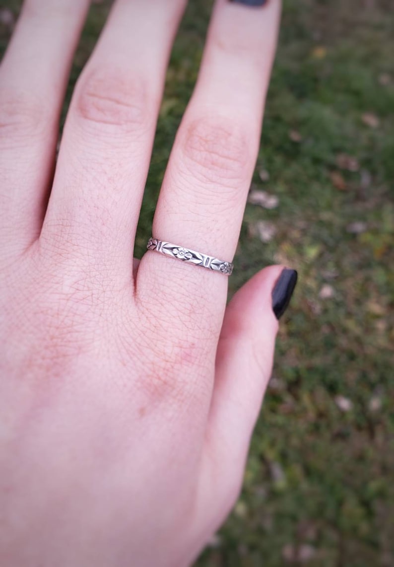 May include: A silver ring with a delicate floral design. The ring is worn on the ring finger of a hand.