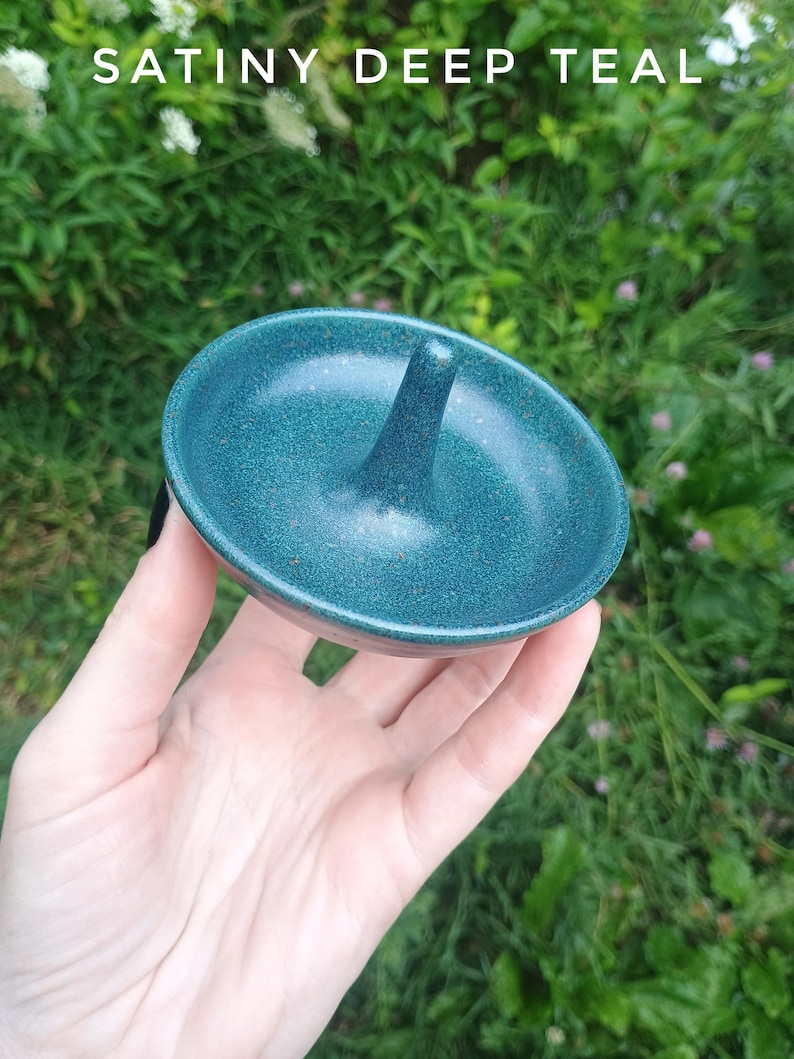 Puede incluir: Un plato de cer&aacute;mica redondo con un cono central para guardar joyas, que presenta un esmalte azul verdoso profundo satinado con una textura sutilmente moteada. El texto SATINY DEEP TEAL aparece impreso en la parte superior de la imagen.