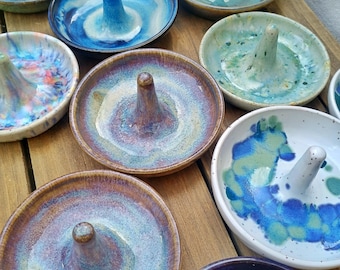 May include: A collection of round ceramic ring dishes, each featuring a central cone for holding jewelry. The dishes display various glossy glaze finishes in shades of blue, green, purple, and white, with some showing speckled or marbled patterns.