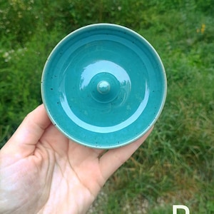 Puede incluir: Un plato de cer&aacute;mica circular de color verde azulado con un acabado liso y brillante con sutiles motas oscuras. El objeto presenta un poste central elevado y redondeado para colocar joyas, y la letra P es visible en la esquina.