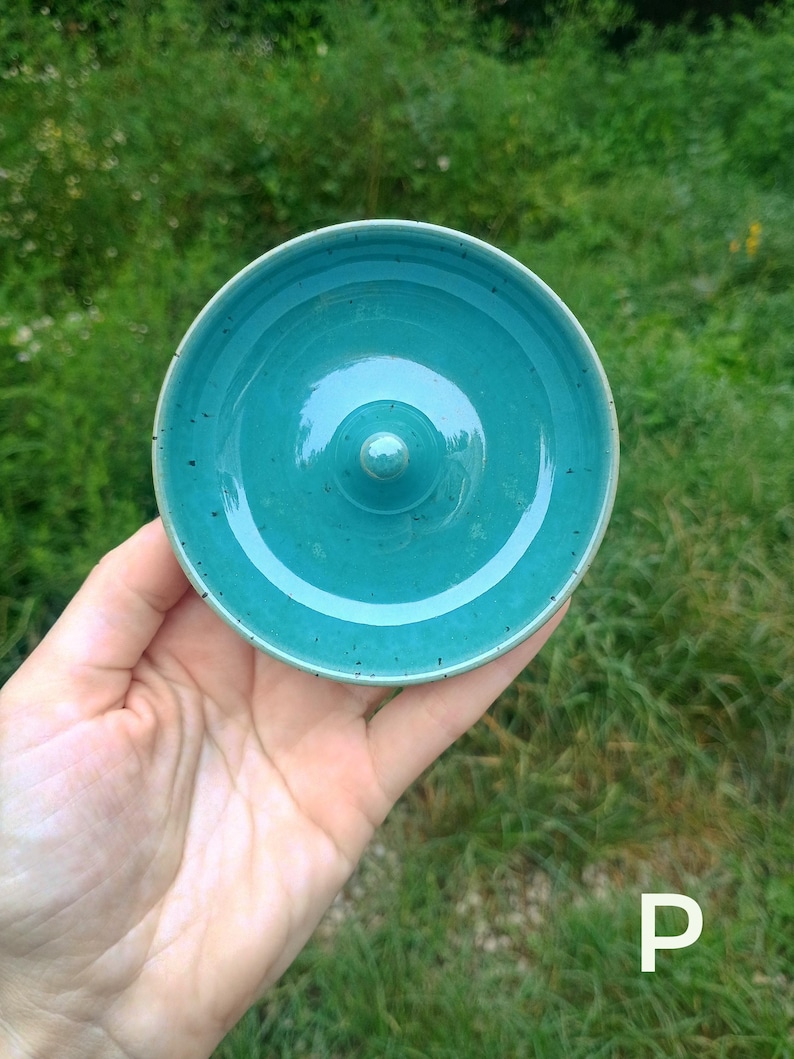 Puede incluir: Un plato de cer&aacute;mica circular de color verde azulado con un acabado liso y brillante con sutiles motas oscuras. El objeto presenta un poste central elevado y redondeado para colocar joyas, y la letra P es visible en la esquina.