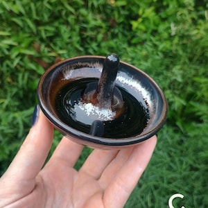 Puede incluir: Un peque&ntilde;o plato de cer&aacute;mica redondo para anillos con un esmalte brillante en tonos marr&oacute;n oscuro y negro, que incluye un poste vertical central. El plato tiene una superficie lisa y reflectante con sutiles motas blancas en la base.