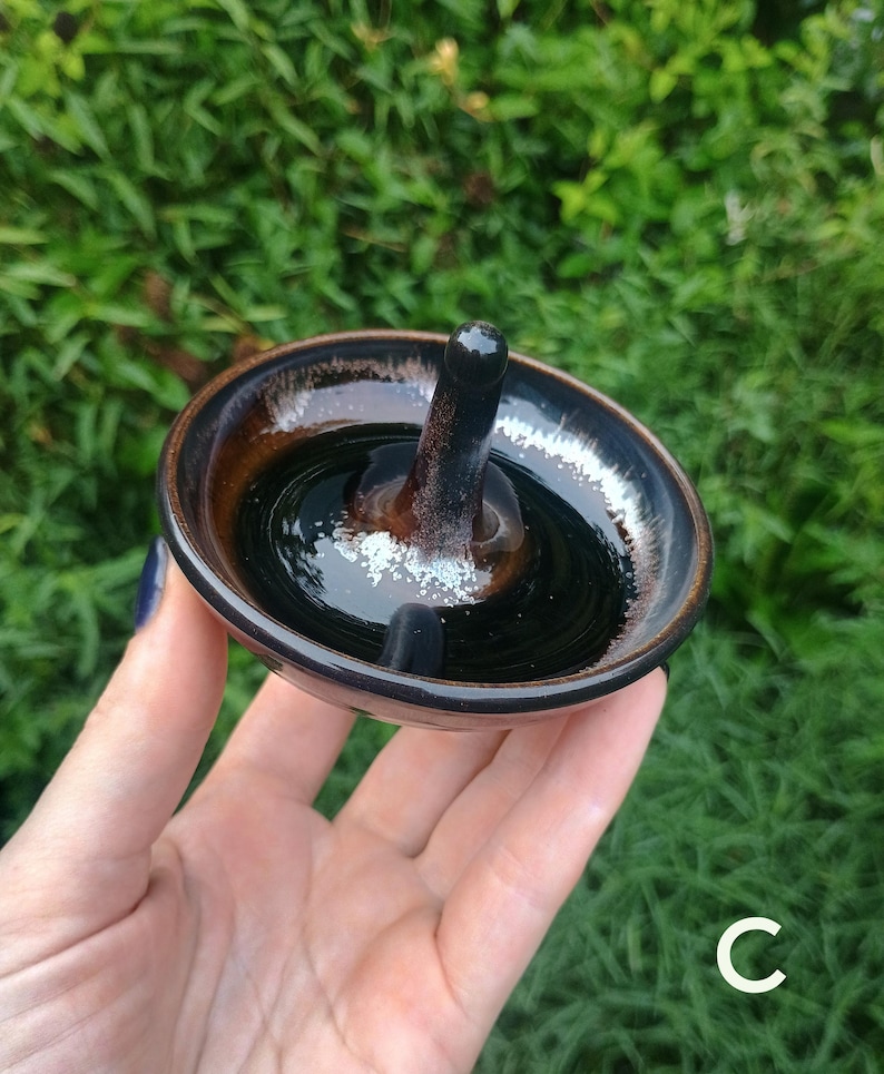 Puede incluir: Un peque&ntilde;o plato de cer&aacute;mica redondo para anillos con un esmalte brillante en tonos marr&oacute;n oscuro y negro, que incluye un poste vertical central. El plato tiene una superficie lisa y reflectante con sutiles motas blancas en la base.