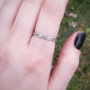 May include: A silver ring with a floral and geometric pattern. The ring is worn on a finger, showcasing its intricate design. The ring's details are visible, highlighting the craftsmanship. The ring is a piece of jewelry.