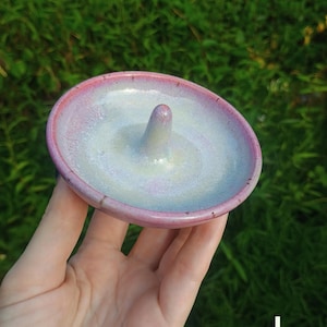 Puede incluir: Un plato de cer&aacute;mica redondo para anillos con acabado brillante, un centro p&aacute;lido e iridiscente que se transforma en un borde rosa vibrante. El plato incluye un poste c&oacute;nico central para sujetar anillos y tiene la letra L en la esquina.