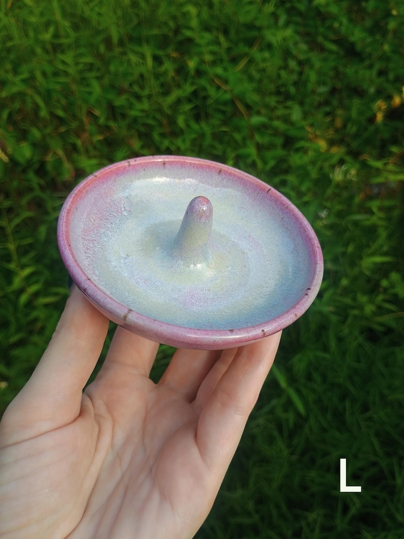 Puede incluir: Un plato de cer&aacute;mica redondo para anillos con acabado brillante, un centro p&aacute;lido e iridiscente que se transforma en un borde rosa vibrante. El plato incluye un poste c&oacute;nico central para sujetar anillos y tiene la letra L en la esquina.