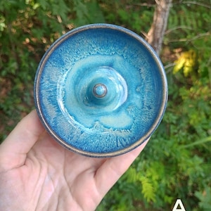Puede incluir: Un plato de cer&aacute;mica redondo para anillos con un esmalte brillante en tonos azul profundo y turquesa, con un cono central. El borde y la base del cono muestran un borde de arcilla marr&oacute;n expuesta. La letra A es visible en la esquina inferior derecha.