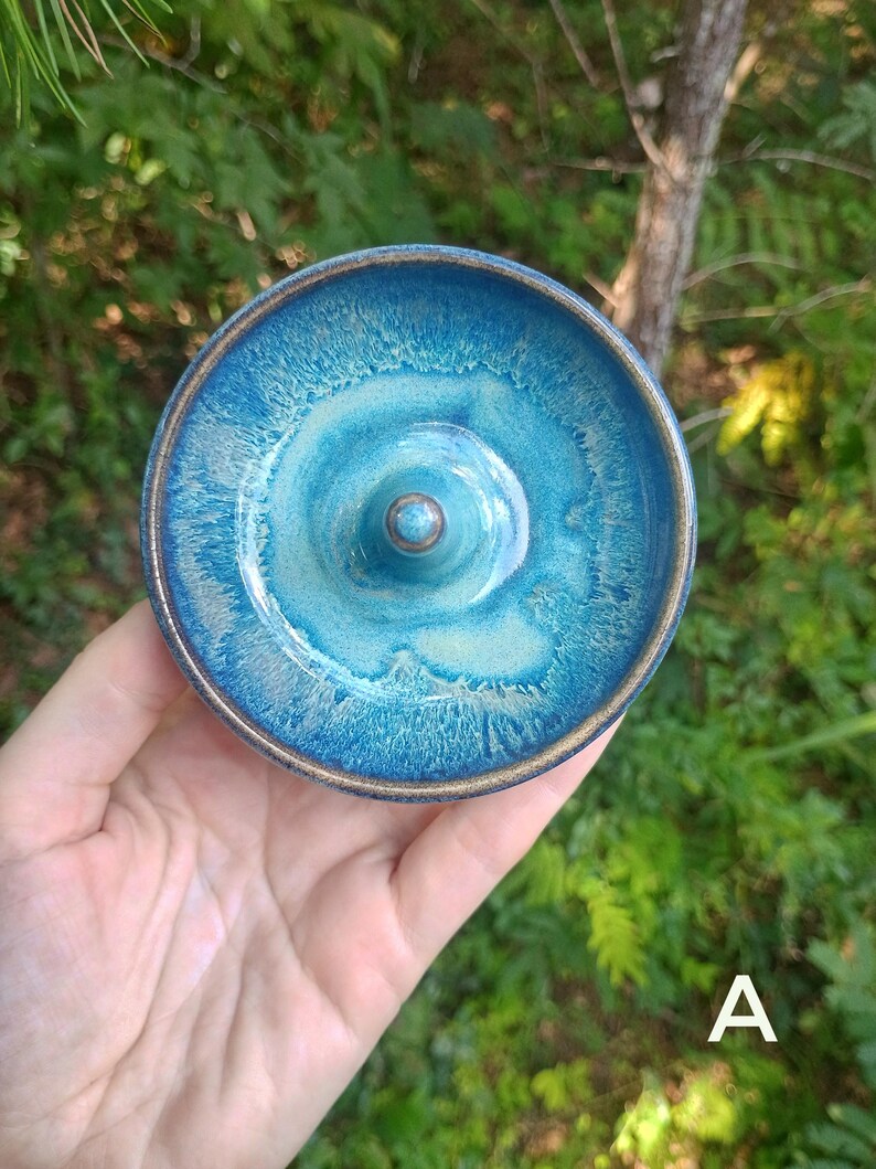 Puede incluir: Un plato de cer&aacute;mica redondo para anillos con un esmalte brillante en tonos azul profundo y turquesa, con un cono central. El borde y la base del cono muestran un borde de arcilla marr&oacute;n expuesta. La letra A es visible en la esquina inferior derecha.