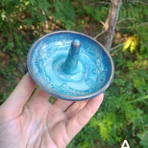 Puede incluir: Un soporte de cer&aacute;mica circular para anillos con un cono central para guardar joyas. La pieza tiene un esmalte brillante con un degradado de azul marino profundo en el borde exterior que transiciona a un azul turquesa vibrante en el centro.