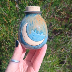 May include: A small, ceramic jar with a cork stopper. The jar is painted with a blue and orange glaze and features a white crescent moon design. The jar is held in a hand against a backdrop of green foliage.