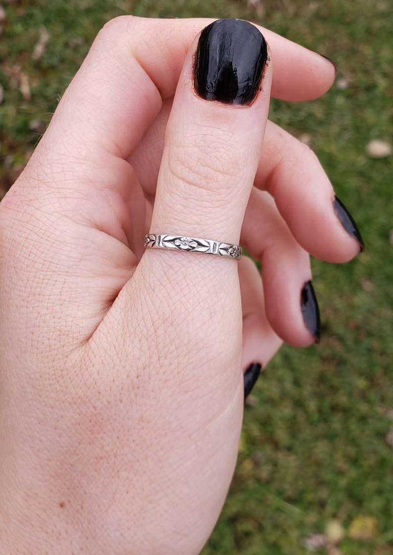 May include: A silver ring with a floral design on the thumb of a hand with black nail polish.