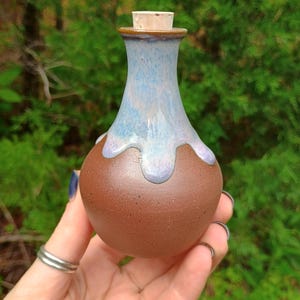 May include: A handcrafted ceramic bottle with a cork stopper. The bottle has a brown base and a light blue and white glazed top. The bottle is held in a hand against a blurred green background.
