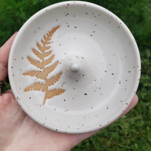 May include: A round ceramic ring dish with a white speckled glaze and a central conical post. The interior surface features a single tan-colored fern leaf imprint. The dish has a smooth, glossy finish and a raised rim, designed for holding jewelry.