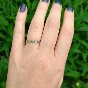 May include: A silver band ring with a delicate geometric design. The ring is worn on the ring finger of a hand with dark blue nail polish.