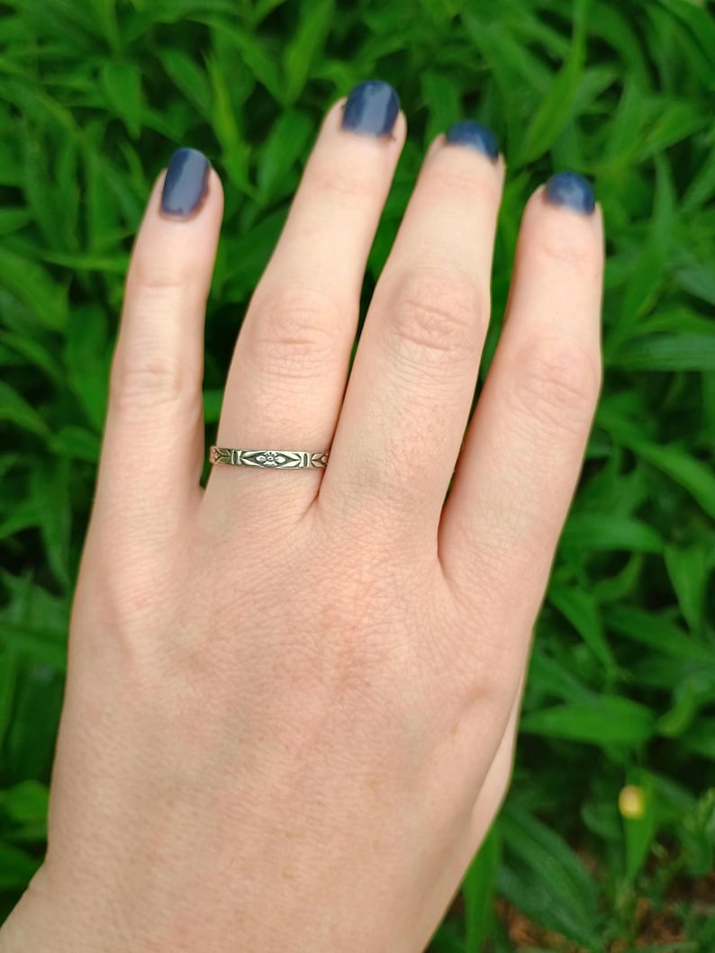 May include: A silver band ring with a delicate geometric design. The ring is worn on the ring finger of a hand with dark blue nail polish.