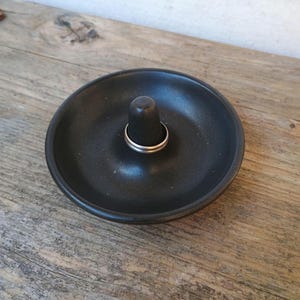 May include: A black ceramic ring holder with a central post. A silver ring rests on the post. The holder is round with a slightly raised edge. The ring holder sits on a wooden surface.