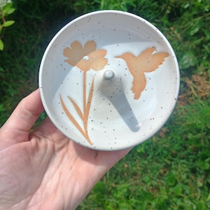 Puede incluir: Un plato de cer&aacute;mica redondo de color blanco roto con acabado moteado y un poste c&oacute;nico central. La superficie interior est&aacute; decorada con una ilustraci&oacute;n de color marr&oacute;n claro de una flor y un colibr&iacute; en vuelo, situados en lados opuestos.