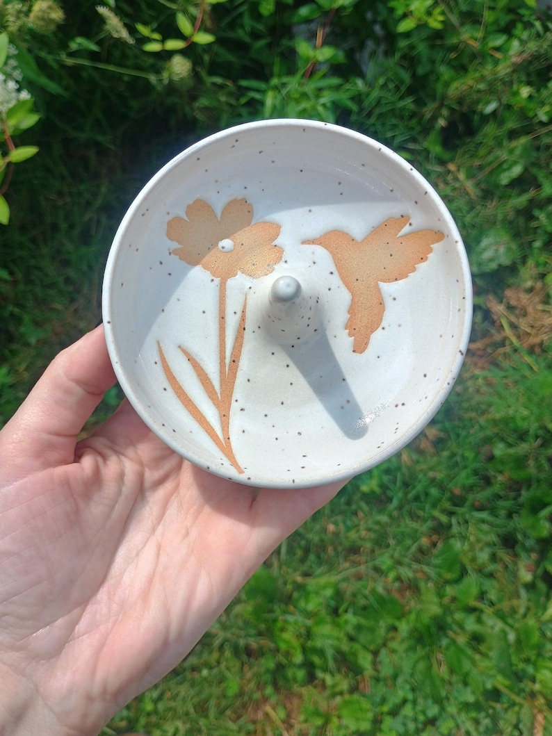 Puede incluir: Un plato de cer&aacute;mica redondo de color blanco roto con acabado moteado y un poste c&oacute;nico central. La superficie interior est&aacute; decorada con una ilustraci&oacute;n de color marr&oacute;n claro de una flor y un colibr&iacute; en vuelo, situados en lados opuestos.