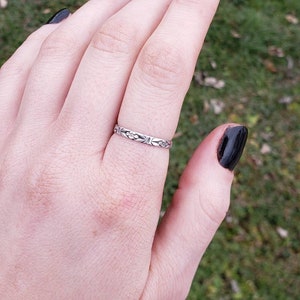 May include: A silver band ring with a floral design. The ring is on a person's finger.