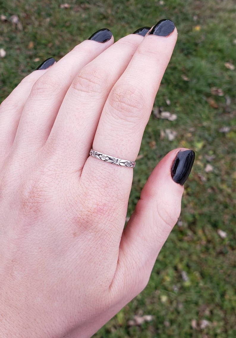 May include: A silver band ring with a floral design. The ring is on a person's finger.