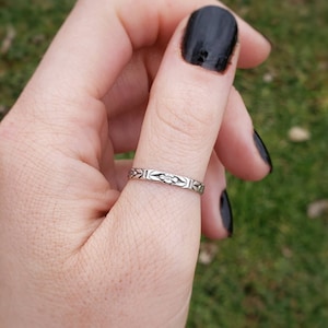 May include: A silver ring with a floral design on the band. The ring is worn on a person's thumb.