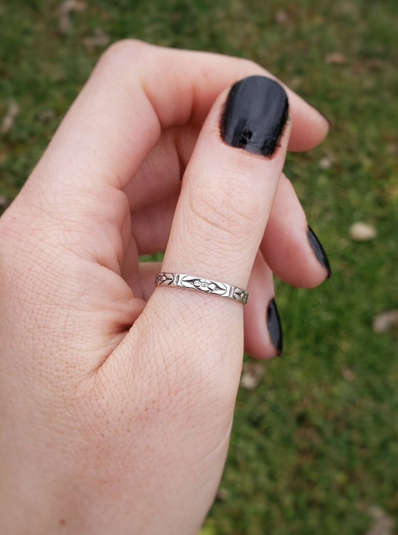 May include: A silver ring with a floral design on the band. The ring is worn on a person's thumb.
