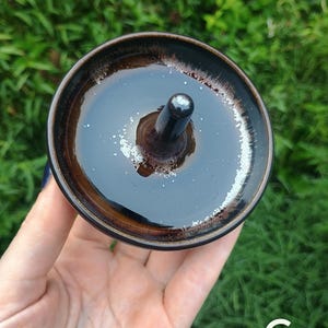 Puede incluir: Un cuenco de cer&aacute;mica circular para lana con un poste central para sujetar el hilo. El cuenco presenta un esmalte marr&oacute;n oscuro brillante con un interior marr&oacute;n m&aacute;s claro y un borde moteado blanco distintivo. La letra C es visible abajo a la derecha.